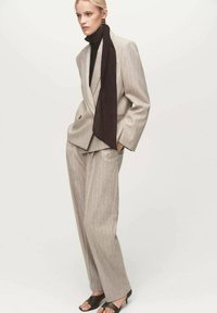Costume beige à rayures avec une veste ajustée et un pantalon ample, associé à une écharpe marron foncé et des talons ouverts noirs.
