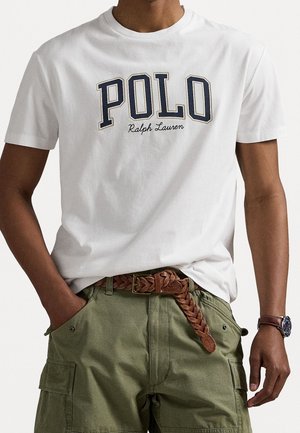 Osoba nosící bílé tričko Polo Ralph Lauren, zelené cargo kalhoty s hnědým pleteným páskem a hodinky na levém zápěstí.