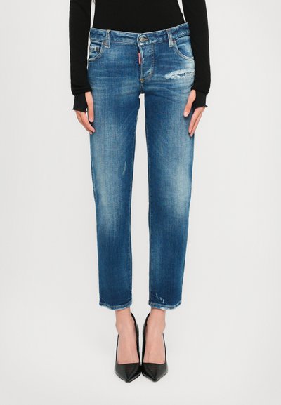 Vrouw in blauwe versleten jeans en een zwart lang mouwtop, staand met de handen langs haar zij, in zwarte puntige hakken, tegen een witte achtergrond.