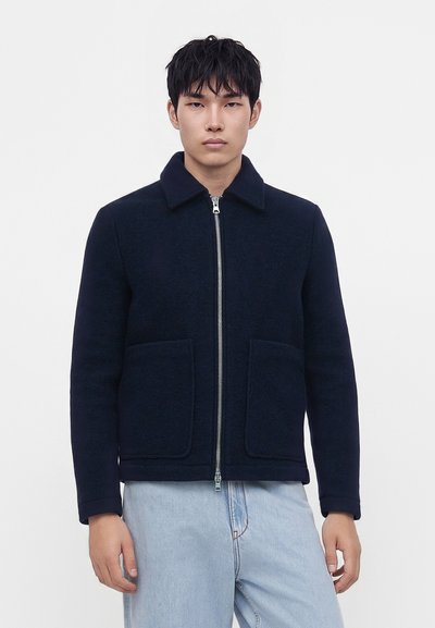 Jeune homme aux cheveux foncés portant une veste bleu marine zippée avec de grandes poches avant et un jean bleu clair sur un fond gris uni.