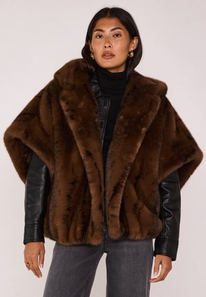 Femme portant un châle en fausse fourrure marron sur une veste en cuir noire, un col roulé noir, un jean gris et des boucles d'oreilles créoles dorées, devant un fond uni.