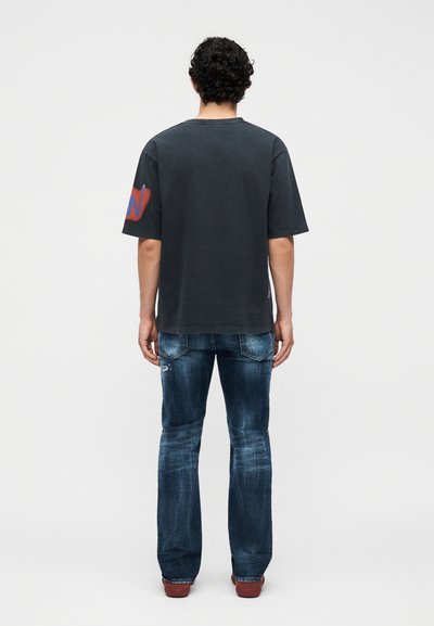 Άνδρας με σγουρά μαλλιά που φοράει μαύρο μπλουζάκι με κόκκινες και μπλε λεπτομέρειες στο μανίκι, μπλε ξεθωριασμένα τζιν και καφέ παπούτσια, στέκεται γυρισμένος από πίσω.