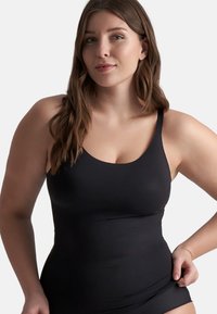 Mujer con cabello largo y castaño que lleva un bañador negro de estilo top, sosteniendo el borde inferior con una mano, fondo neutral.