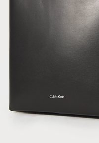 Black leather bag with a smooth texture. Features a prominent white "Calvin Klein" logo at the bottom. Rectangular shape with minimal design.