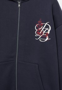 Hanorac cu fermoar de culoare bleumarin, cu un buzunar frontal, prezentând broderie florală roșie și un design cu litere albe pe partea stângă. Material din amestec de bumbac.