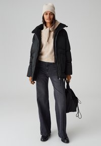 Giacca a vento nera con colletto morbido e peloso, maglione beige, jeans a gamba larga grigio scuro e uno zaino nero. Berretto in maglia strutturata.