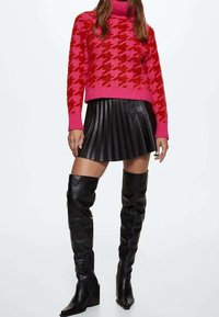 Rosa Strickpullover mit Hahnentrittmuster, schwarzer plissierter Lederrock und knielange schwarze Lederstiefel mit spitzen Zehen.