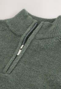 Grøn uldtrøje med høj krave og halvlange lynlås; har en struktureret strik og en skinnende sølvlynlås.