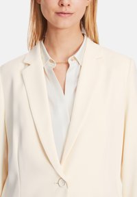 Cream blazer with a structured design, featuring notched lapels and a single button closure. Worn over a light blue shirt with a collar.