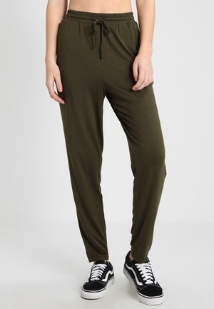 Personne portant un pantalon à cordon vert olive et des baskets noires et blanches, debout devant un fond clair.