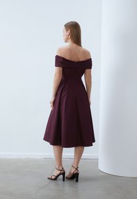 Μπορντό φόρεμα με τους ώμους ακάλυπ�τους, εφαρμοστό μπούστο και φαρδύ φούστα. Λείο υλικό, πλισέ λεπτομέρειες και κρυφός φερμουάρ στην πλάτη.