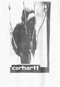 Biała koszulka z nadrukiem w czerni i bieli przedstawiającym postać w kapturze otoczoną cienkimi gałązkami, z logo "carhartt" poniżej.