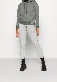 Szary hoodie z kieszenią typu kangurka, z pasującymi ściągaczami na dole i mankietach, zestawiony z jasnoszarymi, lekko przetartymi dżinsami oraz czarnymi butami na sznurówki.