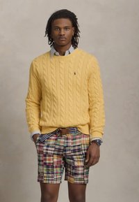 Un pull en maille jaune porté par-dessus une chemise à col bleu clair, associé à un short en tartan multicolore et une ceinture marron. Fond neutre.