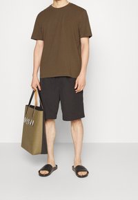 Brun bomulls-t-shirt, svarta shorts, svarta tofflor. Håller en tvåfärgad väska med ett mönstrat accent. Minimalistisk design, avslappnad passform.