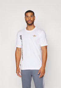 Camiseta blanca de manga corta hecha de algodón, con un pequeño logo dorado en el pecho y texto vertical negro en el lateral.