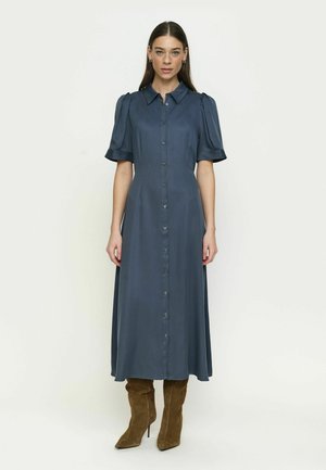 Donna in piedi, indossa un abito midi blu scuro con bottoni, maniche corte e colletto, e stivali marroni in camoscio fino al ginocchio, su sfondo neutro.