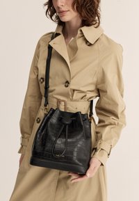 Black leather bucket bag with a textured crocodile pattern, cinch top closure, and adjustable strap, held by a person in a beige trench coat.