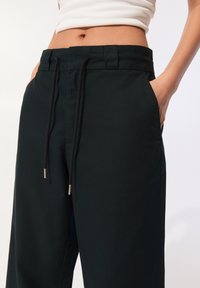 Close-up van een persoon die zwarte drawstringbroek en een witte cropped tanktop draagt, met de handen in de broekzakken.