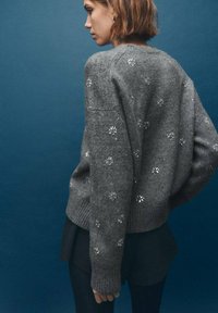 Szary sweter dziergany o luźnym kroju, zdobiony małymi srebrnymi kwiatowymi wzorami, z prążkowanymi mankietami i dołem.