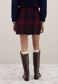 Rückenansicht eines rot-navy karierter Minirock, kombiniert mit kniehohen braunen Lederstiefeln und weißen Socken. Die Textur ist weich und glatt.