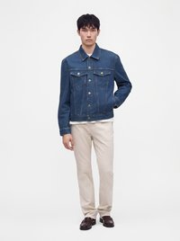 Jeune homme aux cheveux noirs courts portant une veste en jean bleue, une chemise blanche, un pantalon beige et des chaussures marron foncé, debout avec une main dans la poche.