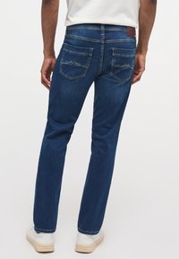 Blugi din denim de culoare albastru închis, cu un design drept, cusături vizibile, două buzunare din spate cu un model în formă de munte, purtați cu încălțăminte albă.