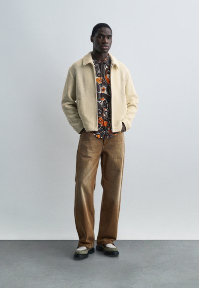 Άνδρας που στέκεται μπροστά σε έναν απλό τοίχο, φορώντας μία κρεμ πλεκτή ζακέτα, ένα πουκάμισο με μοτίβο πορτοκαλί λουλουδιών, καφέ παντελόνι και δίχρωμα παπούτσια.