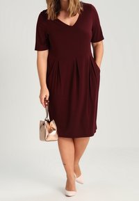 Donna che indossa un vestito bordeaux al ginocchio con maniche corte, porta una borsa metallica e indossa tacchi di colore chiaro, in piedi su uno sfondo semplice.