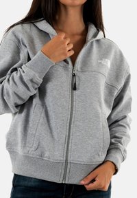 Sudadera gris con cremallera, confeccionada en un tejido suave, que presenta una cremallera frontal, dos bolsillos laterales y puños y dobladillo acanalados. Logotipo exhibido en el pecho.