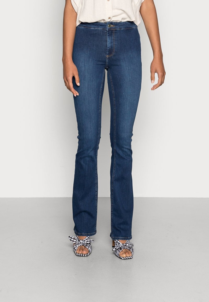 Femme portant un jean slim bootcut bleu foncé, associé à des sandales à talons à carreaux noir et blanc et une chemise blanche à manches courtes.