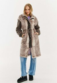Žena v kabátu z umělé kožešiny po kolena s potiskem a stahovací šňůrkou v pase, modrých džínách a černých platformových botách, stojící s rukama v kapsách.