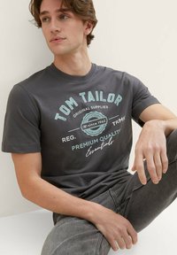 Graues Baumwoll-T-Shirt mit einem bedruckten Design in Hellblau und Weiß, mit dem Text "Tom Tailor" und einem Logo. Kombiniert mit dunkelgrauen Jeans.