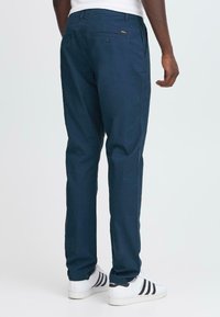 Moški v temno modrih vsakdanjih hlačah in belih Adidas športnih čevljih, stoji s pogledom na hrbtno stran pred enostavno svetlo ozadje.