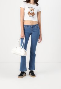 Camiseta corta blanca con un gráfico de osito de peluche, jeans acampanados azules, zapatillas negras con rayas blancas y un bolso blanco.