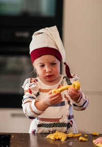 Lapsi päällä valkoinen ja marooni kokinhattu, pitää kädessään keltaista taikinaa ja keskittyy sen muotoon. Raidallinen pitkähihainen paita, jossa on leikkisiä kuvioita.