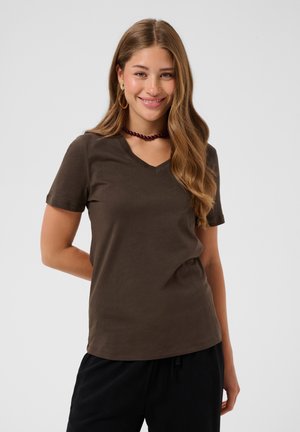 Młoda kobieta z długimi falowanymi włosami uśmiecha się, nosząc brązowy T-shirt z dekoltem w serek, czarne spodnie, złote kolczyki-koła i ciemny naszyjnik z koralików na białym tle.