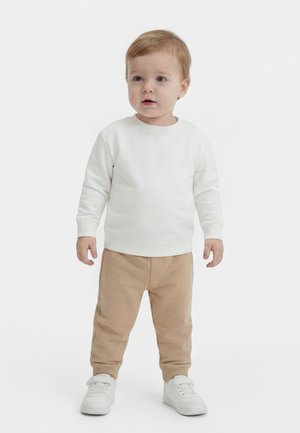 Småbarn står med nøytral uttrykk iført hvit langermet skjorte, beige bukser og hvite joggesko mot ensfarget bakgrunn.