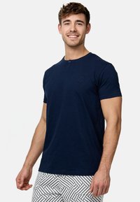 T-shirt bleu marine à manches courtes avec col rond et détail de bouton, présentant un logo de marque discret sur la poitrine. Texture de tissu lisse.