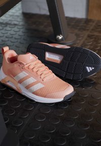 Двойка коралово розови и бели маратонки Adidas върху черен текстуриран под на фитнес зала, като едната подметка е обърната нагоре.