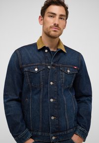 Homme portant une veste en denim bleu foncé avec un col en velours côtelé beige, des boutons et des poches sur la poitrine, se tenant devant un fond uni.