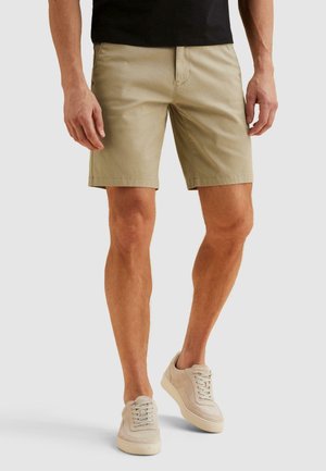 Mann trägt beige knielange Shorts, schwarzes T-Shirt und beige Schnürsneaker, steht vor einem einfarbigen hellen Hintergrund.