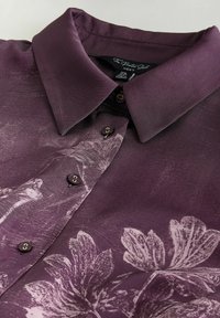 Lilla skjorte med knapper og blomsterprint i lysere lilla. Har klassisk krave og runde mørke knapper, lavet af glat materiale.