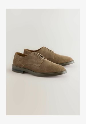 Brązowe zamszowe buty o eleganckim wzornictwie, z zapięciem na sznurowadła i gładką gumową podeszwą. Charakteryzują się spiczastym noskiem i minimalistycznym stylem.