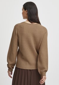Pull marron avec un motif texturé, col rond et manches bouffantes. Porté par-dessus une jupe plissée marron, avec des poignets et un ourlet côtelés.