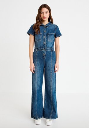 Ung kvinde stående iført en knappet denim-dragt med korte ærmer, brede ben, frontlommer og hvide sneakers.