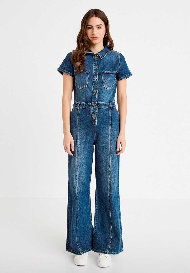 Mujer joven de pie, vestida con un mono de mezclilla abotonado, de mangas cortas, piernas anchas, bolsillos delanteros y zapatillas blancas.