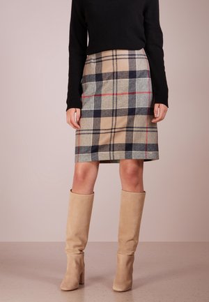 Femme portant une jupe à carreaux beige, noire et rouge jusqu'au genou, un haut à manches longues noir et des bottes à talons beiges montant jusqu'au genou.