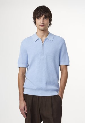 Lyseblå ribbet polo-shirt med lynlåskrave og korte ærmer, parret med brune plisserede bukser. Glat, blødt tekstur.
