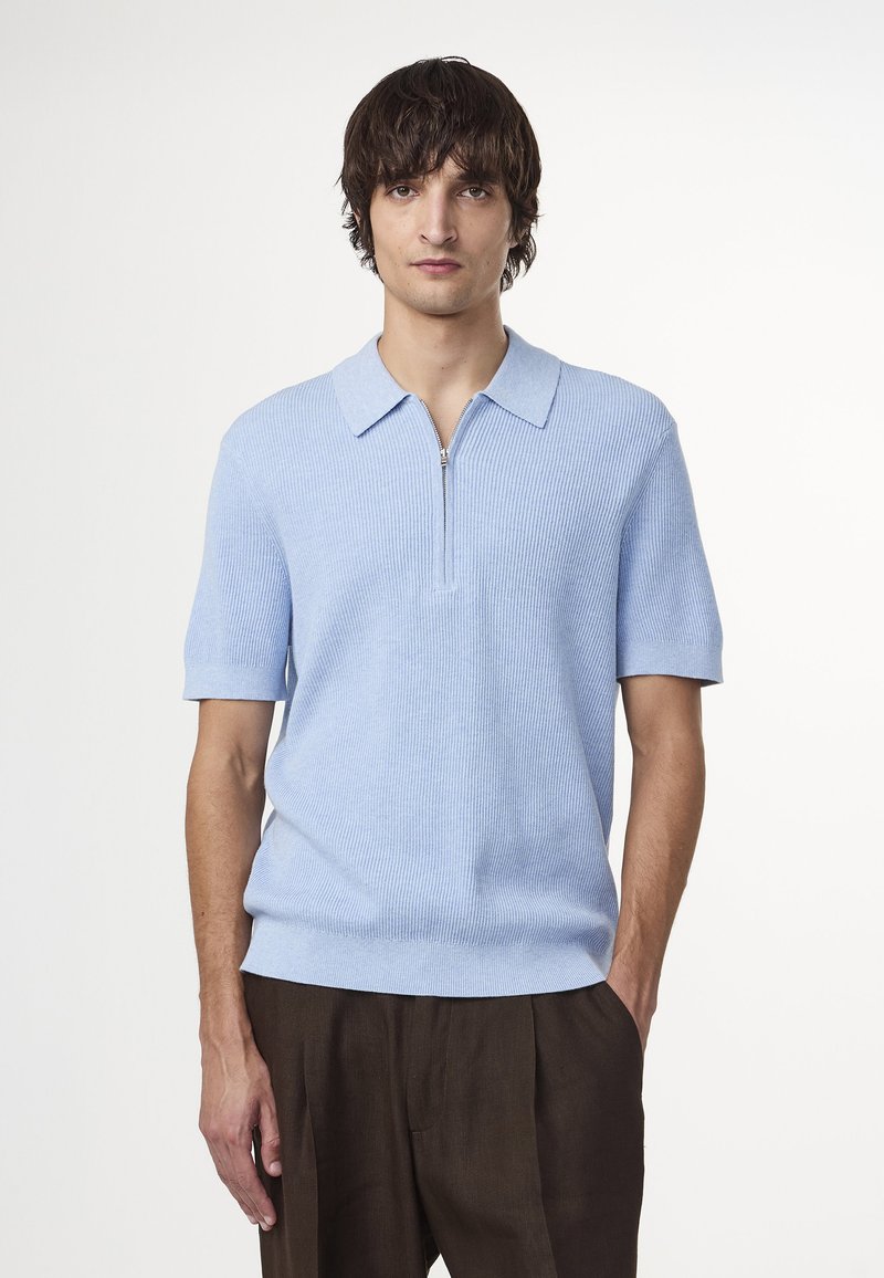 Vaaleansininen ribattua kangasta oleva polo-paita, jossa on vetoketjukaulus ja lyhyet hihat, yhdistettynä ruskeisiin muotoiltuihin housuihin. Suuri, pehmeä rakenne.